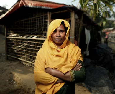 A woman crosses her arms. She is wearing a gold cloth covering and standing in front of a wooden house.